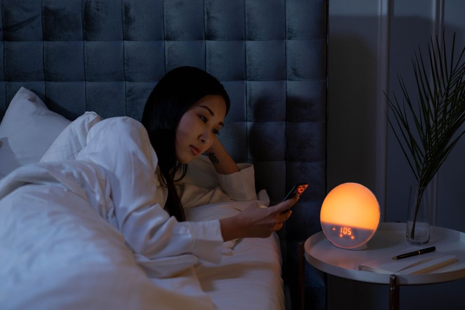 Longevidade — Asian woman in bed using smartphone, illuminated by warm light, indicating insomnia or sleeplessness.
